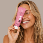 A smiling woman with blonde hair holds a pink tube of Xhilaration XO2 Professional Perfume against her face, set against a solid taupe background.